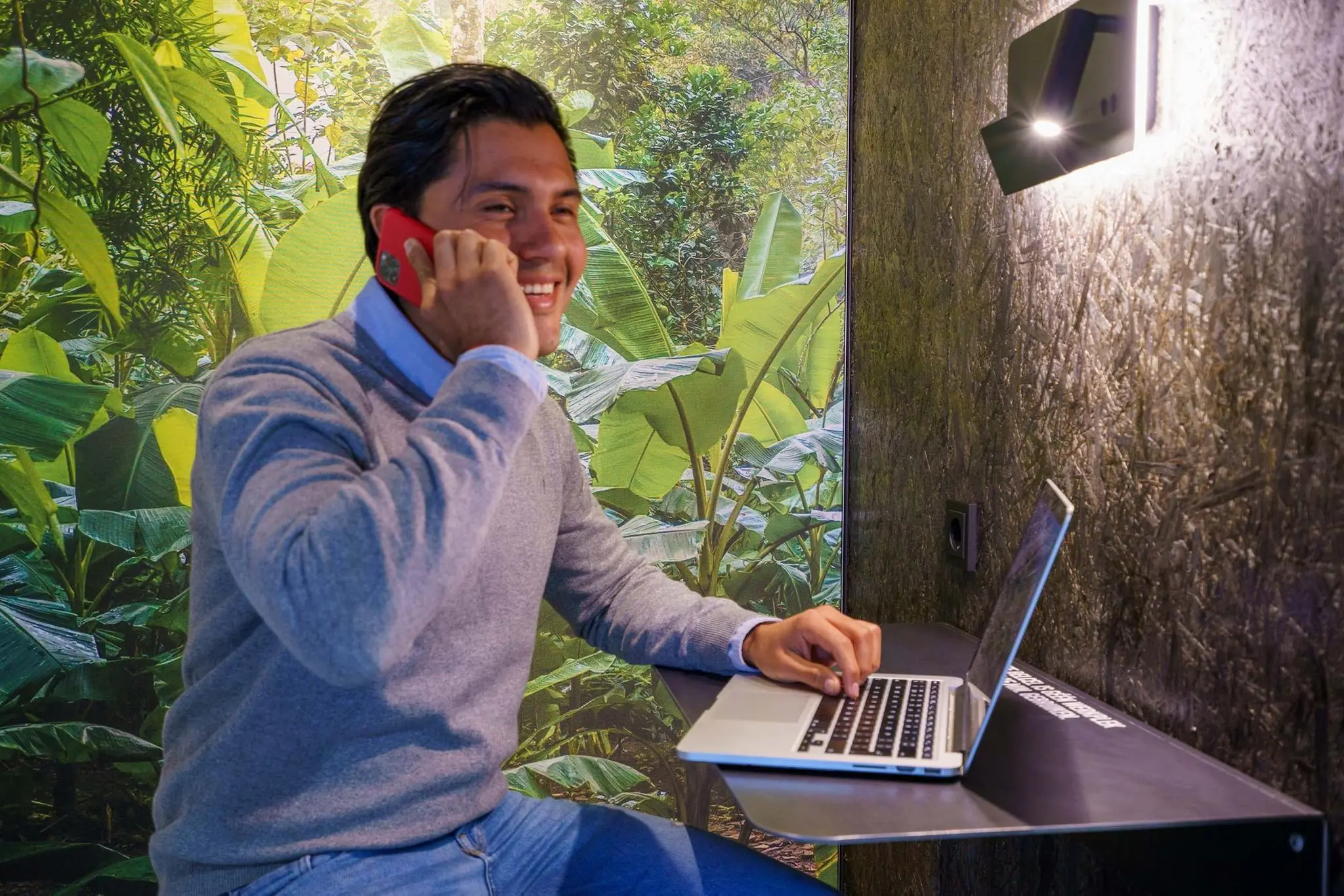 Man in een belcel aan de Baarnsche Dijk voert een telefoongesprek terwijl hij op een laptop werkt, met een tropische plantenmuur op de achtergrond.